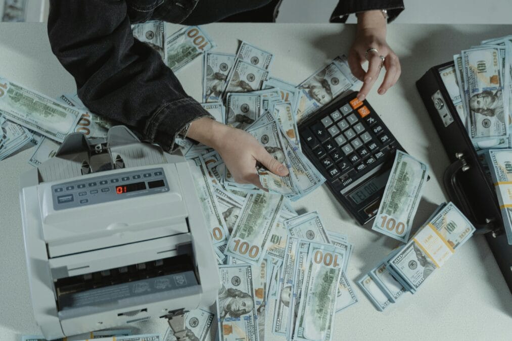Hands counting 100 dollar bills using a calculator and money counter on a table.