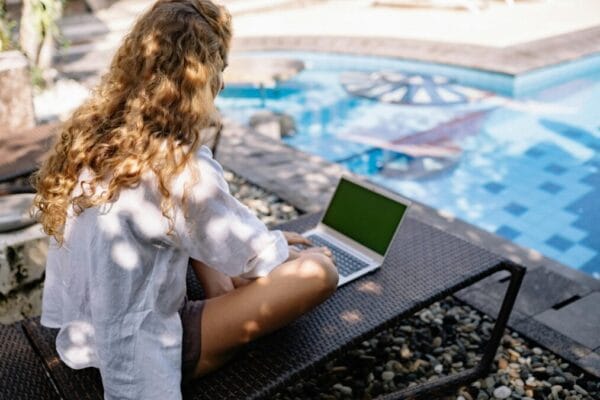 Woman with curly hair working on a laptop outdoors by a pool during summer, embodying a digital nomad lifestyle.